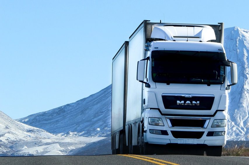Weißer Lkw fährt auf einer Straße mit schneebedeckten Bergen im Hintergrund.